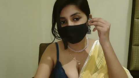 Media: Video of a young woman with medium-dark skin, dark hair, wearing a blue top, black mask, and gold sari, adjusting her hair, indoors.