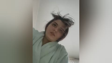 Media: A video of a young woman with short, messy dark hair, wearing a light blue bathrobe, standing in a dimly lit room with white walls. The image has a slight blur.