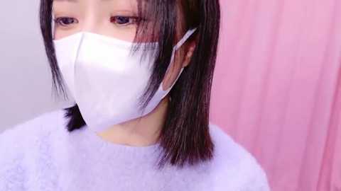 Media: Video of a woman with straight black hair and a white face mask, wearing a white sweater, standing against a soft pink background.