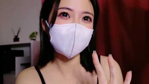 Media: Video of an Asian woman with fair skin, straight black hair, wearing a white surgical mask, black tank top, and red background. She has a neutral expression and is holding her hand up.