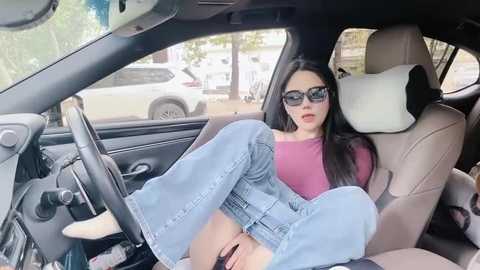 Media: Video of a young woman with long black hair, wearing oversized sunglasses, a red top, and blue flared jeans, sitting provocatively on the passenger seat of a car, legs spread, revealing her vagina.