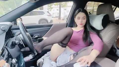 Media: Video of an Asian woman with long black hair, wearing a pink top and black stockings, sitting in a car with a car seat in the background.