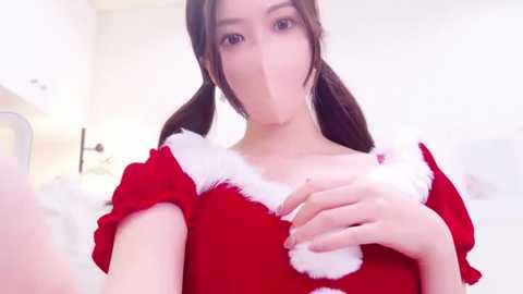 Media: Video of an East Asian woman with long brown hair in a red Santa outfit with white fur trim, holding her chest, in a bright, white room with blurred background.