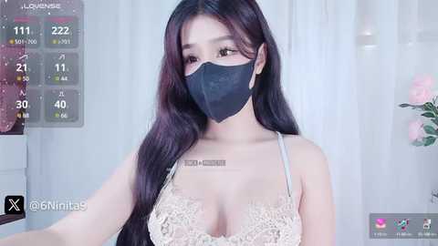 Media: A young Asian woman with long black hair, fair skin, and large breasts, wearing a black face mask and a beige lace bra, stands in a bright, modern room with white curtains and a pink flower vase.