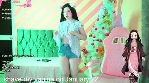 Media: A video of a young Asian woman with long black hair, wearing a white polka-dotted blouse and denim shorts, standing next to a decorated Christmas tree in a pink room. A life-sized Naruto figurine is on the right.