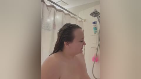 Media: Video of a fair-skinned woman with wet, shoulder-length brown hair, showering in a white-tiled bathroom. She has a medium build and is topless. The background includes a showerhead, soap dispensers, and a partially visible towel.