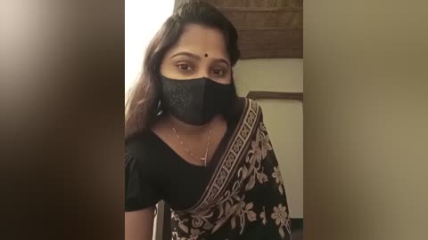 Media: A video of a South Asian woman with medium skin tone, wearing a black mask, black top, and floral sari with a gold border. She has straight, dark hair and a dot on her forehead. Indoor setting with a wooden ceiling and beige walls.