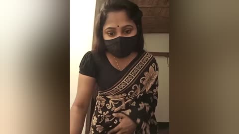 Media: Video of a South Asian woman in a black mask, black blouse, and ornate sari with gold embroidery, standing indoors with a beige wall and wooden door in the background.