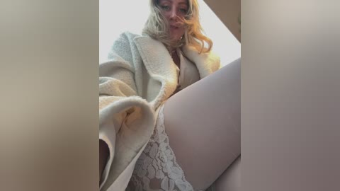 Media: A video of a blonde woman in a white lace bra, wearing a beige cardigan, with her legs spread, taken from a low angle.