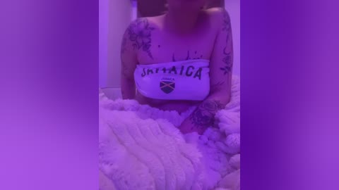 Media: A video of a topless, tattooed woman with light skin, holding a towel with \"Jamaica\" written on it, lying in a bed covered in fluffy white bedding. The room is bathed in purple light.