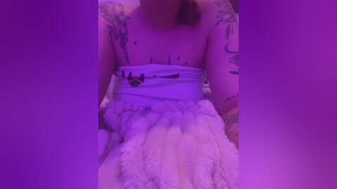 Media: Video of a woman with light skin and tattoos, wearing a white, fluffy dress, lying on a bed under purple lighting.