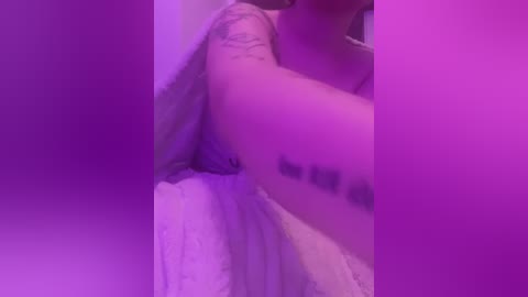 Media: Video of a person with fair skin, visible tattoo, and purple lighting. They are partially clothed, lying on a white bed, creating a sensual, intimate atmosphere.