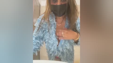 Media: Video of a woman with long, straight brown hair, wearing a blue fuzzy robe, black mask, and a gold bracelet. She is taking a selfie in a bathroom with beige walls.