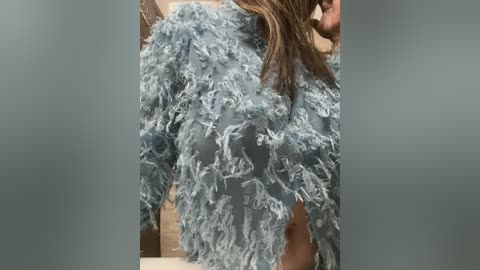 Media: Video of a person wearing a blue, fluffy, feather-textured jacket. The jacket has a soft, voluminous texture with long sleeves. The background is blurred, focusing on the jacket.