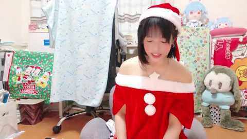 Media: A video of an Asian woman in a red Santa outfit with white trim, kneeling on a wooden floor surrounded by plush toys and festive wrapping paper.