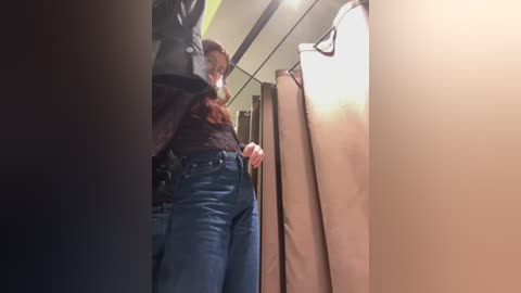 Media: Video of a woman in a dark sweater and jeans, adjusting a hanger in a clothing store's changing room, with beige and white garments hanging nearby.