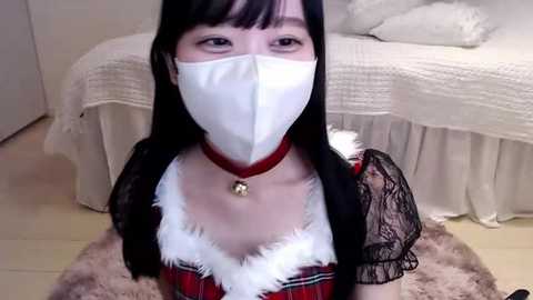 Media: Video of an Asian woman with long black hair, wearing a white mask, red plaid dress, and black lace sleeves, kneeling on a soft, fluffy rug in a softly lit bedroom with a white bed and curtains.