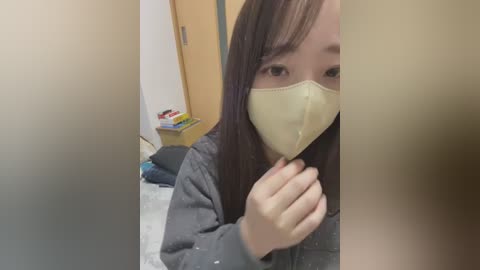 Media: A video of an East Asian woman with long dark hair and light skin, wearing a yellow face mask, a gray sweatshirt, and sitting on a bed. The background shows a wooden wardrobe and scattered clothes.