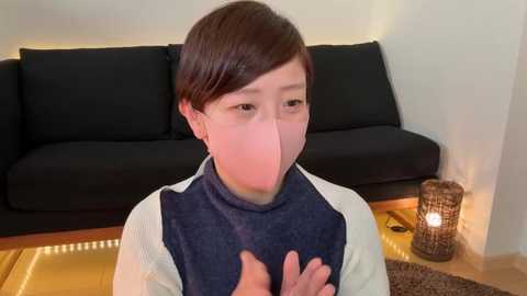 Media: Video of an Asian woman with short, dark hair, wearing a navy blue and white long-sleeve shirt, seated indoors. She is covering her mouth with her hands, and a black couch is in the background.