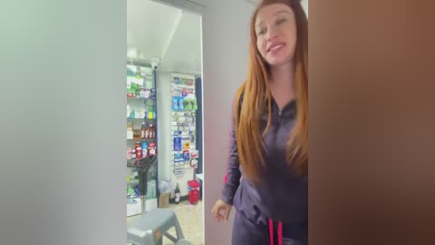 Video of a smiling, long-haired redhead in a dark tracksuit, standing in a brightly lit, modern pharmacy with shelves of colorful products.