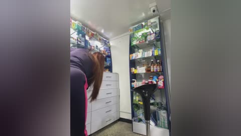 Media: Video of a small, cramped kitchen with a person bent over, facing away, wearing a dark jacket. Refrigerator and cabinets filled with various food items.