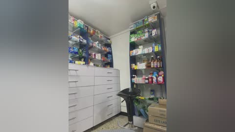 Media: A video of a small, well-organized pharmacy with white shelves filled with various medications and toiletries. A white cabinet with multiple drawers is on the left, and a black stool is centrally positioned.