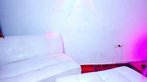 Media: Video of a minimalist, modern living room with a white leather sofa against a white wall, illuminated by soft, purple-pink LED lights. A small, white end table with a phone charger is visible on the right.