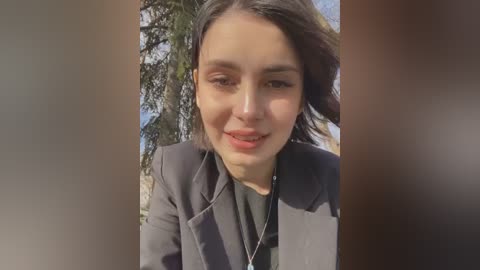 Media: Video of a young woman with light brown skin, shoulder-length dark hair, and a light gray blazer, smiling outdoors with a blurred pine tree background.