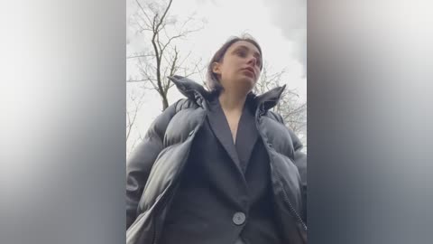 Media: Video of a young woman with fair skin, wearing a black puffy jacket, standing outdoors in a cloudy, leafless environment. Her expression is neutral, and she gazes upward.