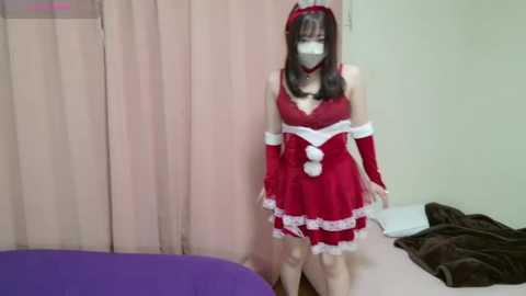 Media: Video of an East Asian woman in a red Santa-themed cosplay outfit with white trim, wearing a face mask, standing indoors with beige curtains and a purple couch in the background.