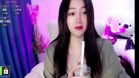 Media: A video of an East Asian woman with long black hair, wearing a green top, holding a drink, in a bedroom with purple lighting, plush toys, and a potted plant.