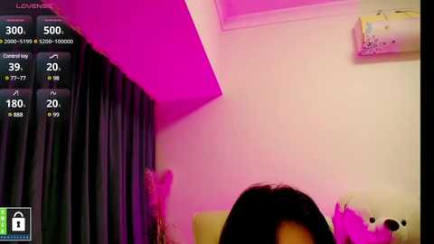 A video of a woman's head in a room with pink lighting, dark curtains, and a white ceiling fan. The background includes a TV screen displaying a weather forecast.