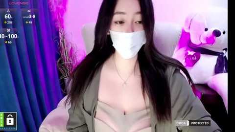 Media: Video of a young Asian woman with long black hair, wearing a white surgical mask, olive green jacket, and beige top, seated in a chair with purple and pink lighting, plush toys in the background.