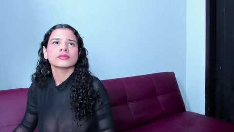 Media: Video of a young woman with curly black hair, wearing a sheer black top, sitting on a maroon leather couch against a light blue wall.