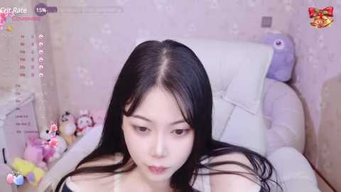 Media: Video of a young Asian woman with long black hair, wearing a white top, sitting on a white couch in a pastel-colored room. The background features plush toys and a calendar.