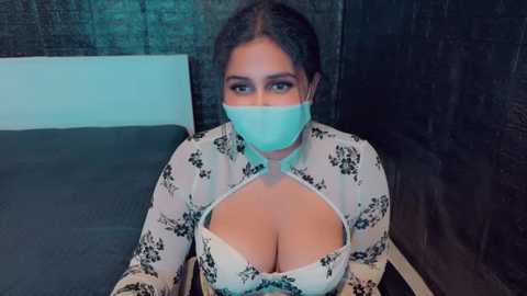 Media: Video of a woman with light brown skin, wearing a white floral blouse revealing her large breasts, a blue surgical mask, and sitting on a bed with dark bedding.