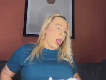 Media: Video of a fair-skinned, blonde woman with a round face and large breasts, wearing a blue t-shirt, sticking her tongue out, against a red wall with a framed abstract painting.