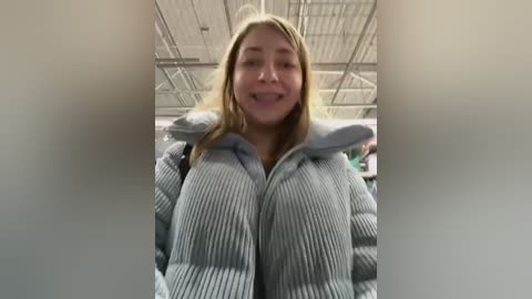 Media: Video of a smiling young woman with light skin and long blonde hair, wearing a light grey puffer jacket, standing inside a large industrial building with a high ceiling and metal beams.