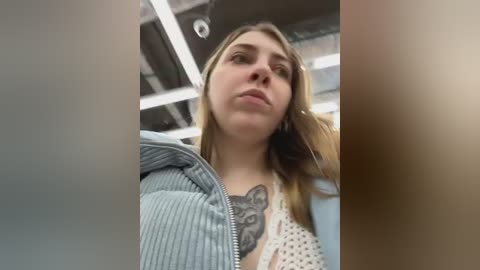 Media: Video of a young woman with long, light brown hair, wearing a white knit sweater with a large black cat tattoo on her chest. She's indoors with industrial ceiling lights and exposed beams in the background.