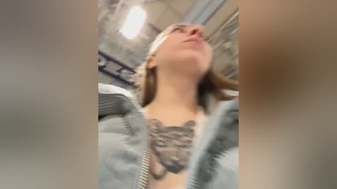 Media: Video of a woman with fair skin, wearing a gray hoodie and a black lace bralette, showing off a detailed chest tattoo. She has light brown hair and is seen from an upward angle in a dimly lit room with visible ceiling lights.