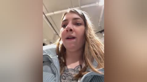 Media: A video of a young woman with light skin and long, straight, brown hair, wearing a light blue plaid jacket over a gray sweater with a detailed pattern. She appears to be indoors with a neutral background.