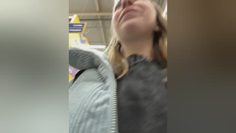 Media: Video of a woman with shoulder-length blonde hair, wearing a gray sweater, laughing and tilting her head back in a grocery store aisle. Background shows yellow and blue signs and industrial ceiling.