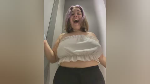 Media: Video of a young Asian woman with shoulder-length light brown hair, wearing a white strapless crop top and black pants, standing in a narrow hallway, mouth open, hands raised.