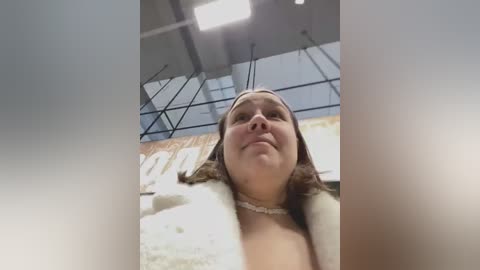 Media: Video of a young woman with light skin and brown hair, wearing a white coat and necklace, standing in an industrial-looking room with high ceilings and metal beams.