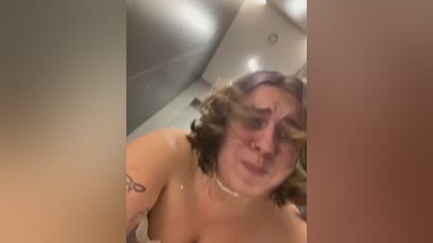 Media: Video of a woman with curly hair, wearing a choker necklace, sitting in a bathtub with visible tattoos on her shoulder, water splashing, blurred background.