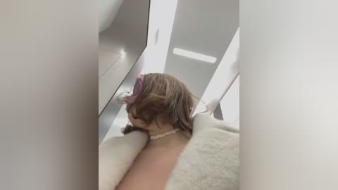 Media: Video of a woman with shoulder-length brown hair, wearing a white fur coat, lying on a white fur rug, viewed from the side through a mirrored closet door.