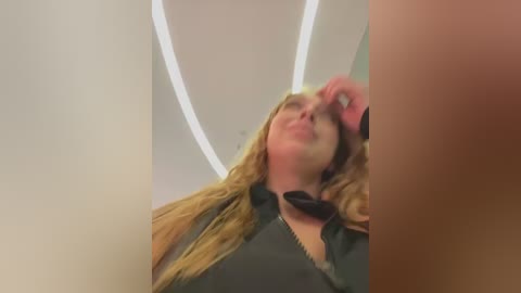 Media: Video of a blonde woman with long hair, wearing a black shirt, lying on a massage table with a white, circular light fixture above her head.