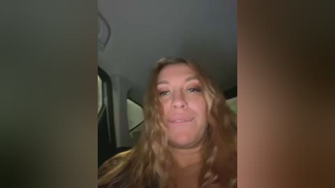 Media: Video of a smiling Caucasian woman with long, wavy blonde hair, wearing a brown jacket, seated in a dimly lit car interior.