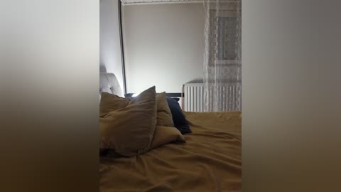 Media: A video of a dimly lit bedroom featuring a neatly made bed with beige sheets and two pillows, a radiator, and sheer curtains partially covering a window.