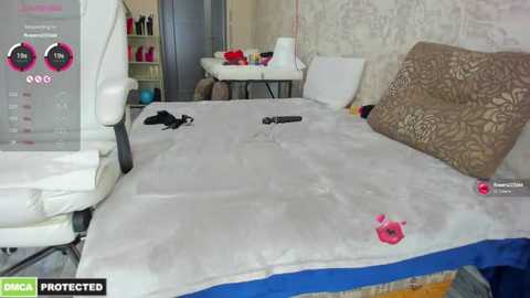 Media: Video of a messy bedroom with a bed covered in white sheets and a blue blanket, scattered clothes, and a pink flower on the floor. Background shows a desk with a chair, a chair with a pink cushion, and a mirror.